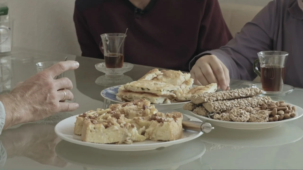 A Gift from God (2019) movie still: medium — A glass dining table holds several white plates containing cakes, bread, nuts, and biscuits. Two people…
