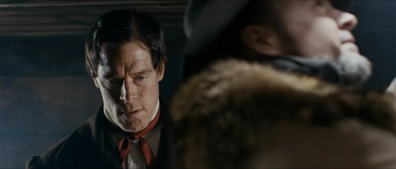 Out of Liberty (2019) movie still: medium — A dimly lit 19th-century wooden interior space with dark walls and shadows. A man in a dark coat looks…