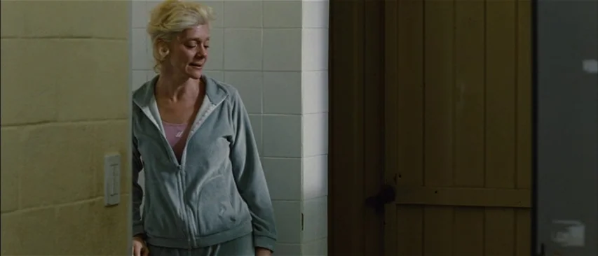 The Headless Woman (2008) movie still: medium — A utilitarian locker room features tiled walls and a wooden door. A woman in a grey velour tracksuit…