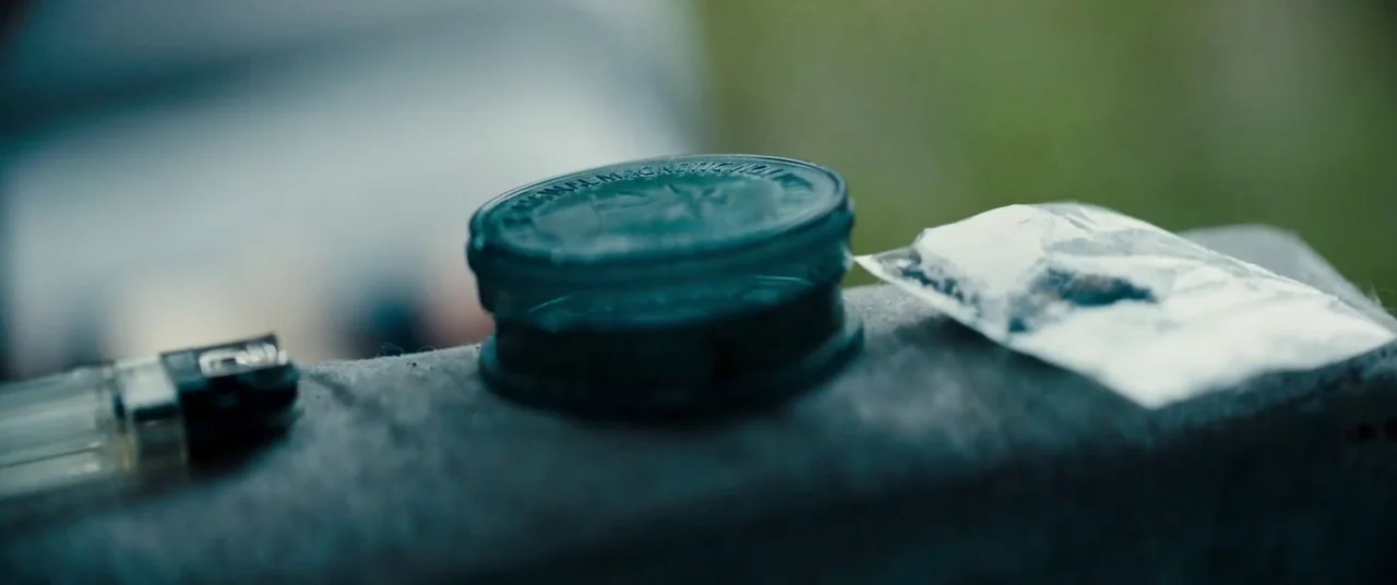 Silver Haze (2023) movie still: closeup — A dark green circular container, a clear plastic baggie, and a lighter rest on a textured concrete…