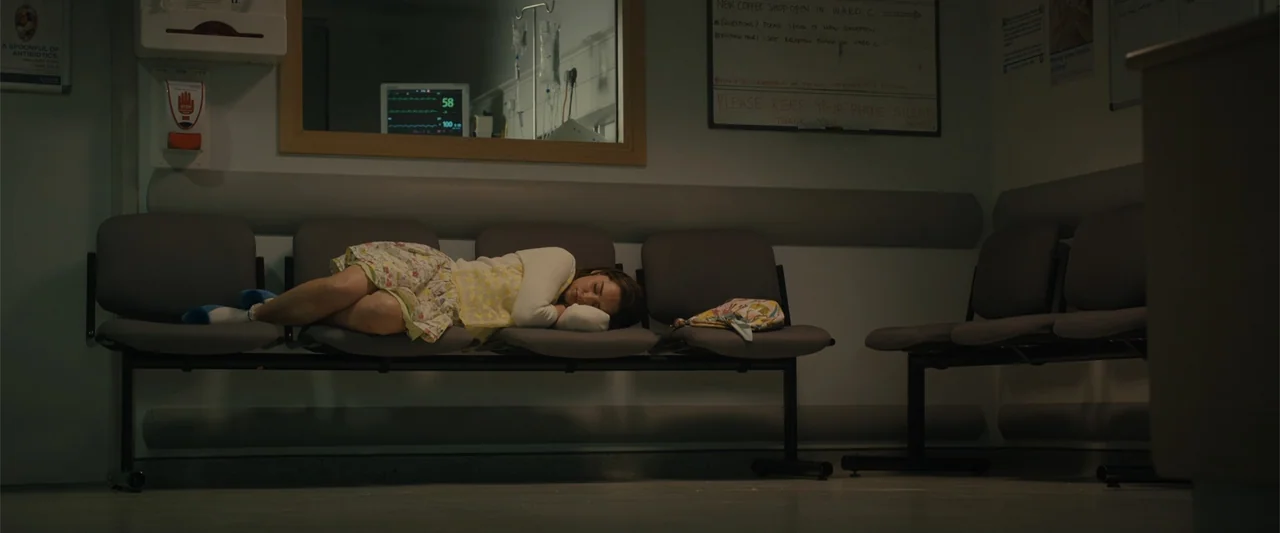 Me Before You (2016) movie still: medium — A quiet hospital waiting area with dark plastic chairs and clinical bulletin boards. A woman in a floral…