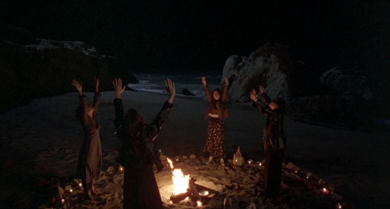 The Craft (1996) movie still: wide — A secluded sandy cove at night with large dark rock formations framing the ocean. Four young women stand in a…