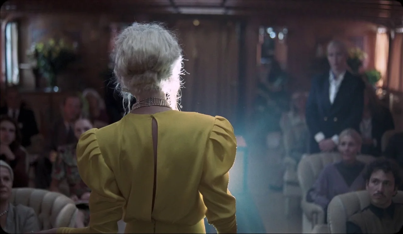 La Chimera (2023) movie still: medium — A dark interior of a vintage passenger carriage with wood paneled walls. A woman in a yellow dress stands…
