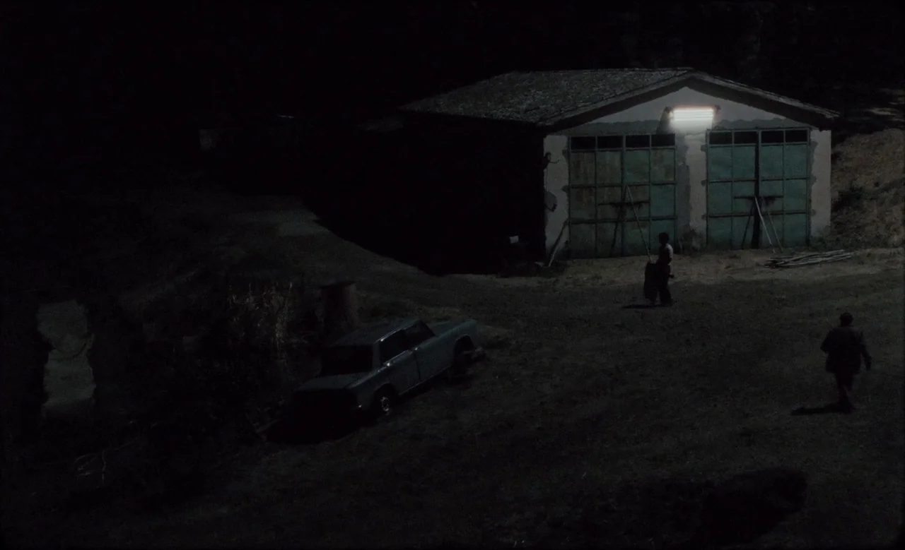 Happy as Lazzaro (2018) movie still: wide — A dark night setting features a small garage with green metal doors and a light. Two men walk across the…