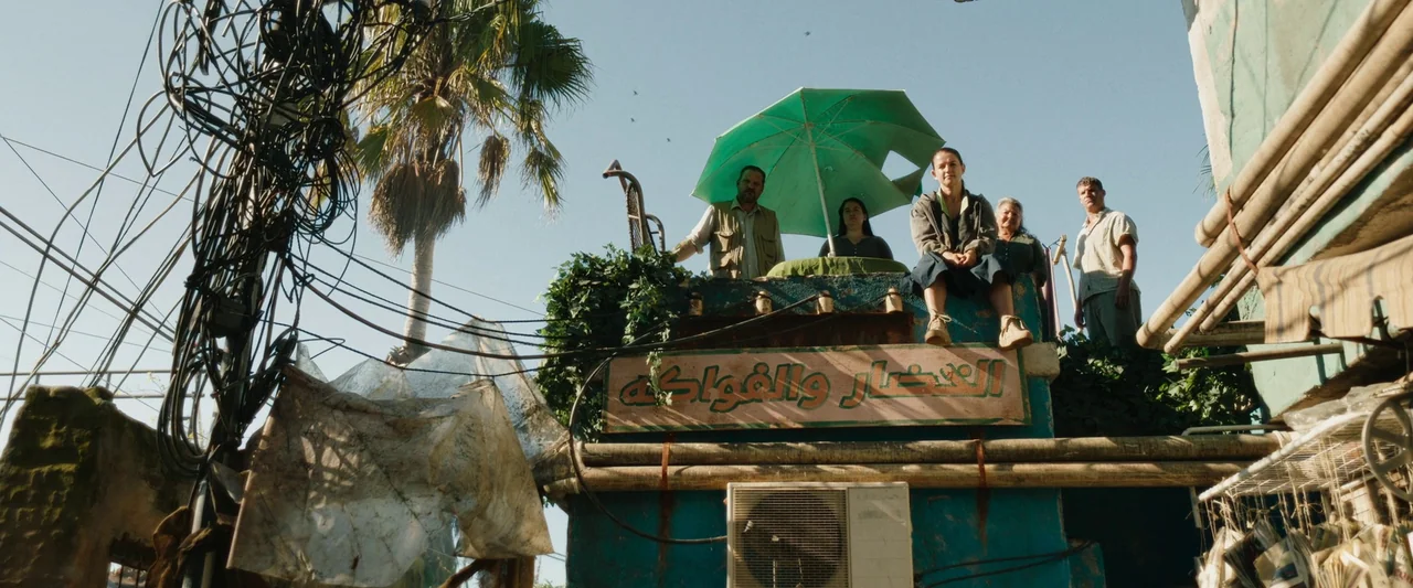 Boy Kills World (2023) movie still: wide — A group of people stands on the rooftop of a weathered, painted masonry building. A sign with Arabic…