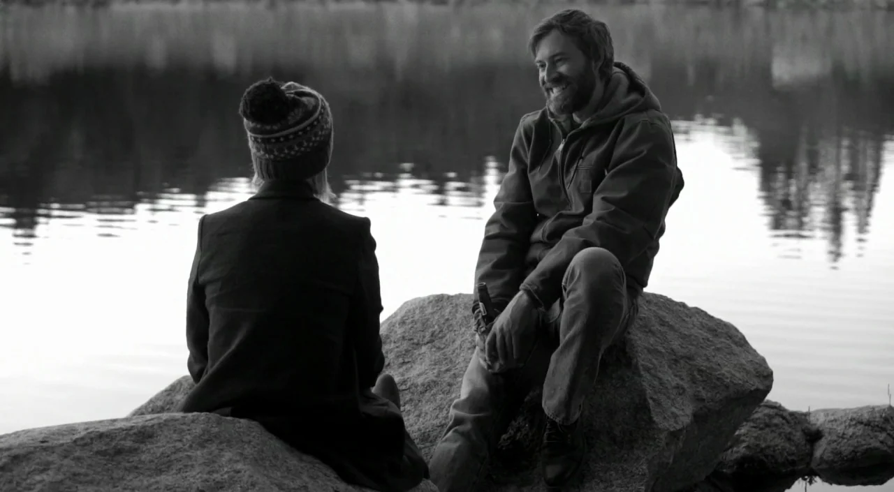 Blue Jay (2016) movie still: medium — A man and woman sit on a large rock at the edge of a calm lake. The man is smiling at the woman while holding…