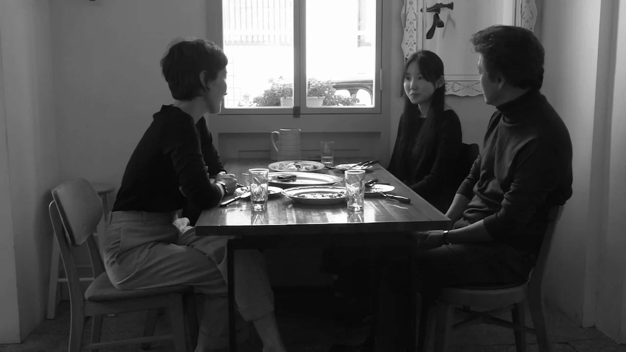Walk Up (2022) movie still: medium — Three people sit at a wooden dining table in a room with white walls. They are having a meal near a window that…