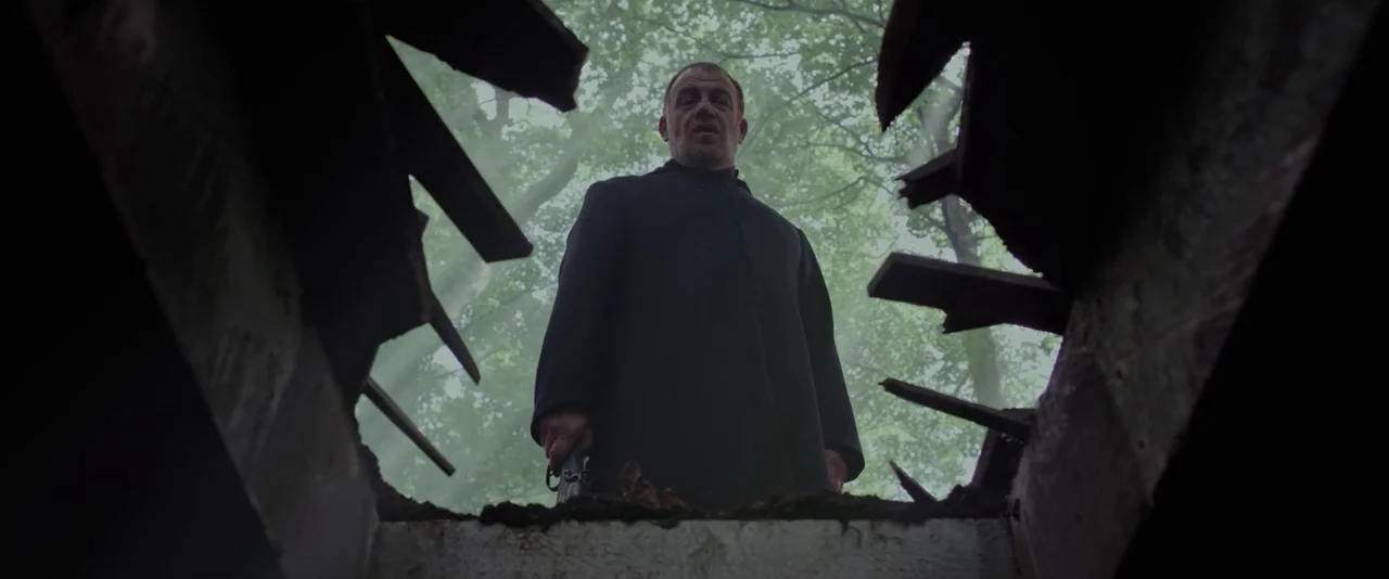 Apostle (2018) movie still: medium — A man stands at the edge of a deep, earth-walled trench within a forest. He looks downward directly into the…