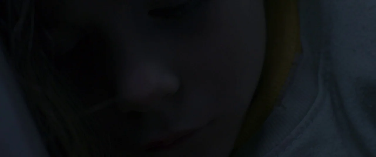 Room (2015) movie still: closeup — A dimly lit interior space with a person partially obscured by darkness. The subject rests in a position…
