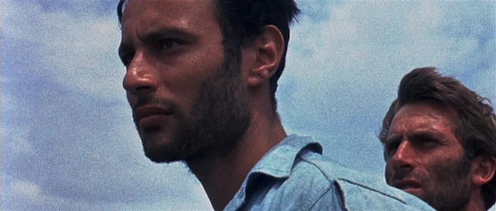 The Intruder (2004) movie still: medium — Two men stand outdoors under a cloudy sky. The camera focuses on the side profile of the man in the…