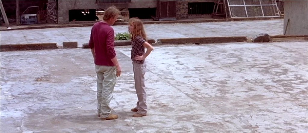 White Material (2009) movie still: fullbody — A wide, cracked concrete lot sits between industrial buildings with metal fencing in back. A man and a…