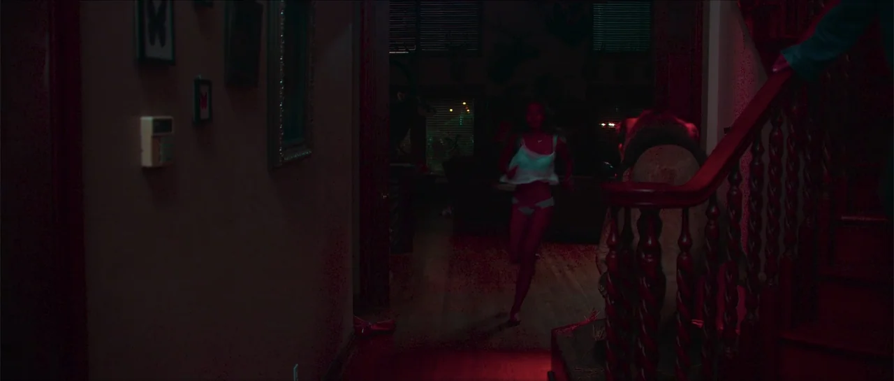 68 Kill (2017) movie still: medium — A dimly lit indoor hallway with a wooden staircase and dark walls. A woman in light clothing runs toward the…