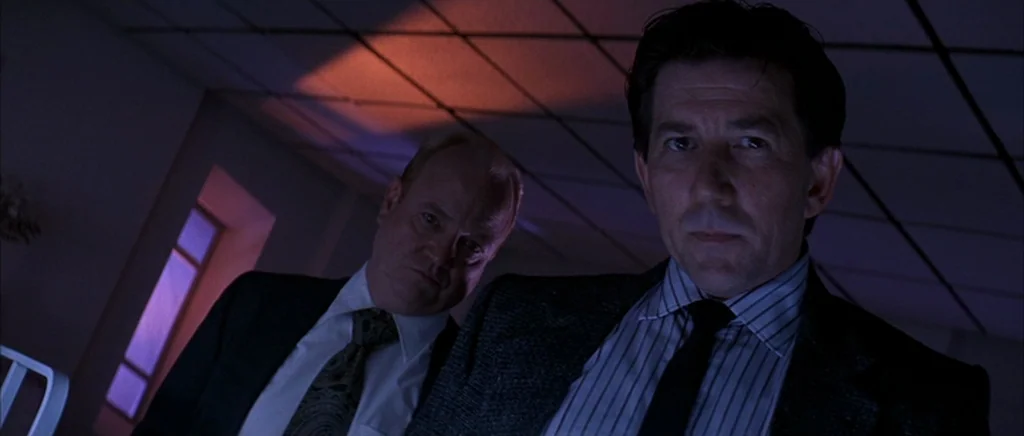The Underneath (1995) movie still: medium — Two men stand over the camera in a darkened room with a tiled ceiling. They gaze downward with stoic…