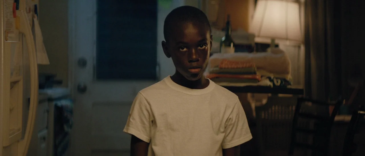 Moonlight (2016) movie still: medium — A dimly lit room contains a white refrigerator and a lamp-lit surface with towels. A young boy in a white…