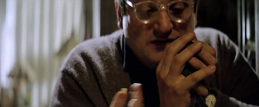 Anguish (1987) movie still: closeup — A man wearing clear-framed glasses sits indoors in a brightly lit room. He holds a small, rounded object in…
