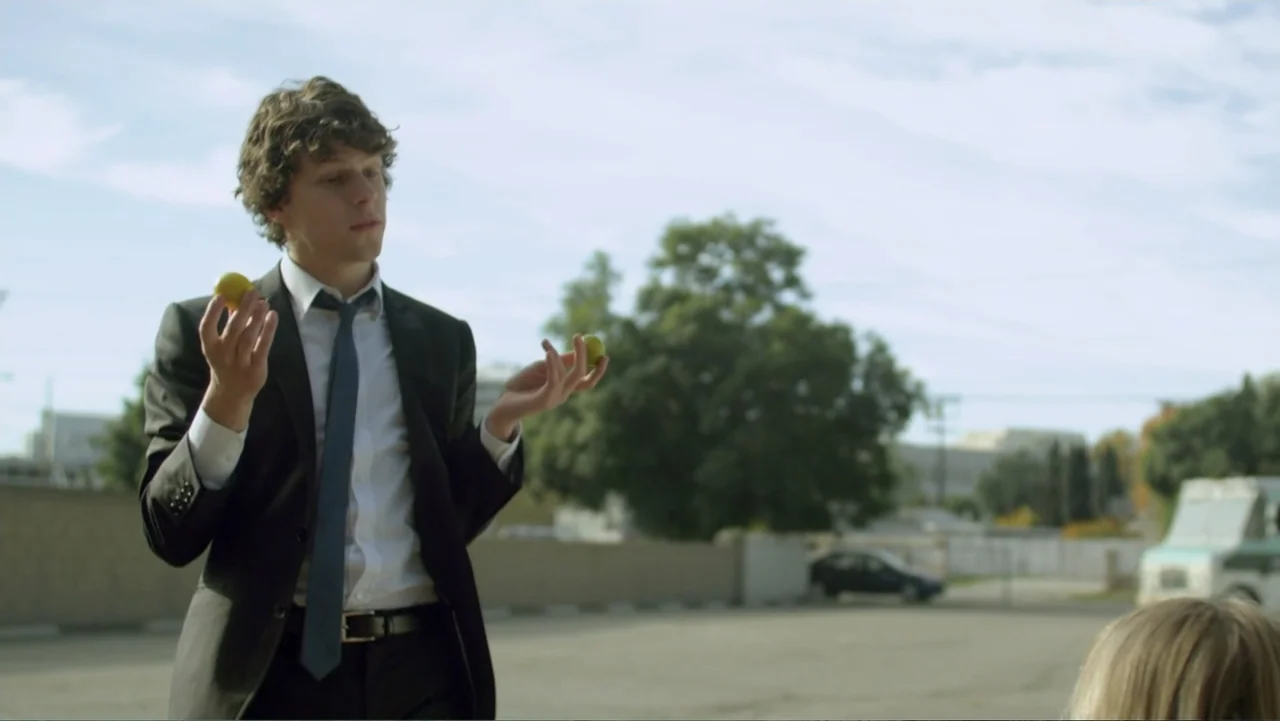 Free Samples (2013) movie still: medium — An asphalt parking lot serves as the setting with a blurred car and trees behind. A man in a business suit…