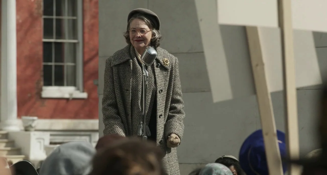 Motherless Brooklyn (2019) movie still: medium — A woman stands behind a silver vintage microphone in front of a stone building. She faces forward…