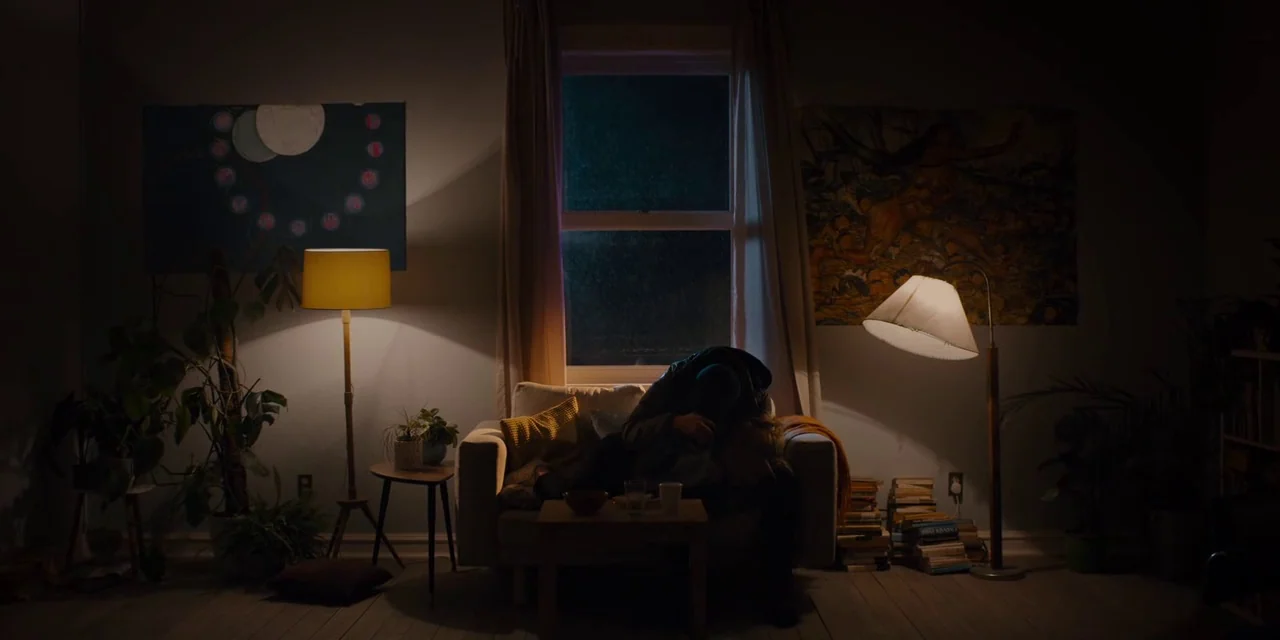 Midsommar (2019) movie still: wide — A dimly lit living room at night contains a sofa beneath a dark window. A person sits on the sofa with their…