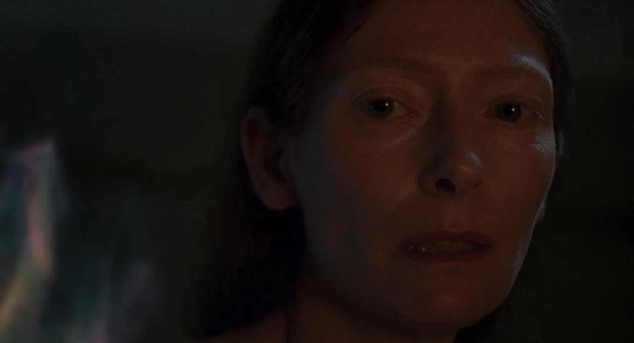 Suspiria (2018) movie still: closeup — A dark, minimally lit environment provides focus on a woman's face. The subject stares ahead with a steady,…