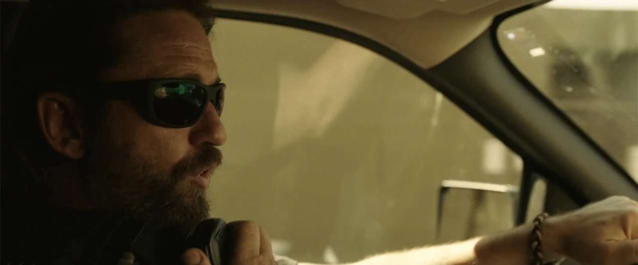 Den of Thieves (2018) movie still: closeup — A man drives a vehicle while using a handheld radio microphone inside the cabin. Sunlight filters…