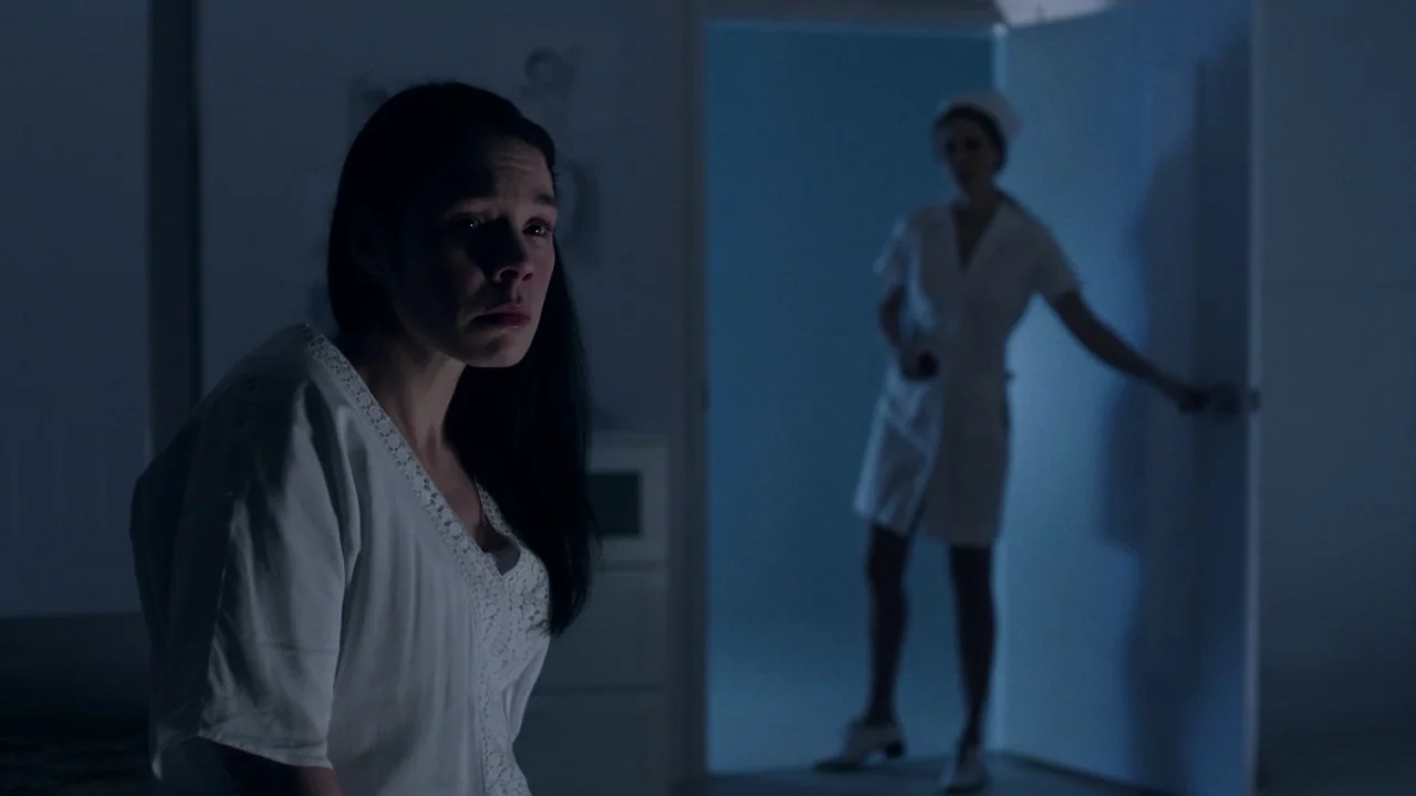 Cult of Chucky (2017) movie still: medium — A dimly lit room with blue shadows and a patient sitting in the foreground. A nurse wearing a…