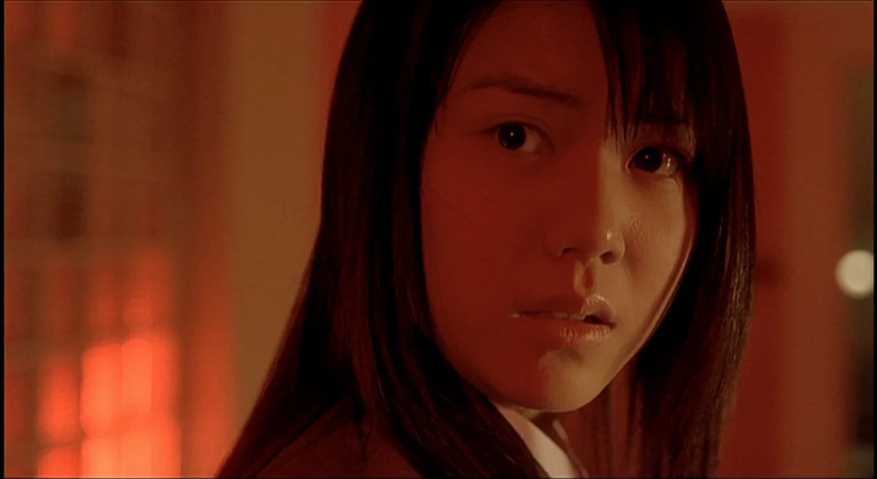Voice (2005) movie still: closeup — A dark indoor space is filled with intense red lighting from a side source. A young woman with long dark hair…