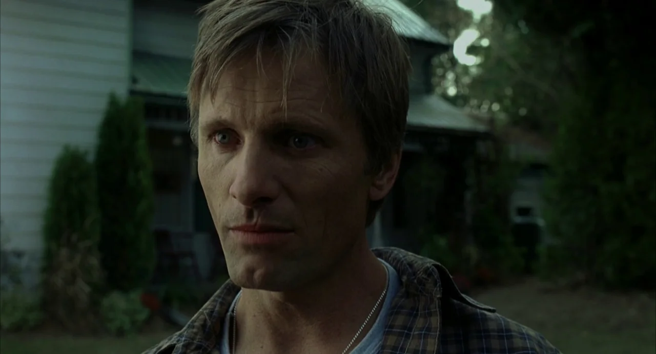 A History of Violence (2005) movie still: closeup — A man stands before a residential building with dark foliage and wooden architecture. The…