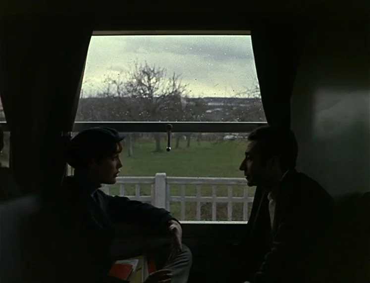 La Chinoise (1967) movie still: medium — Two individuals sit on opposite sides of a train cabin facing each other. Outside the window, a green field…