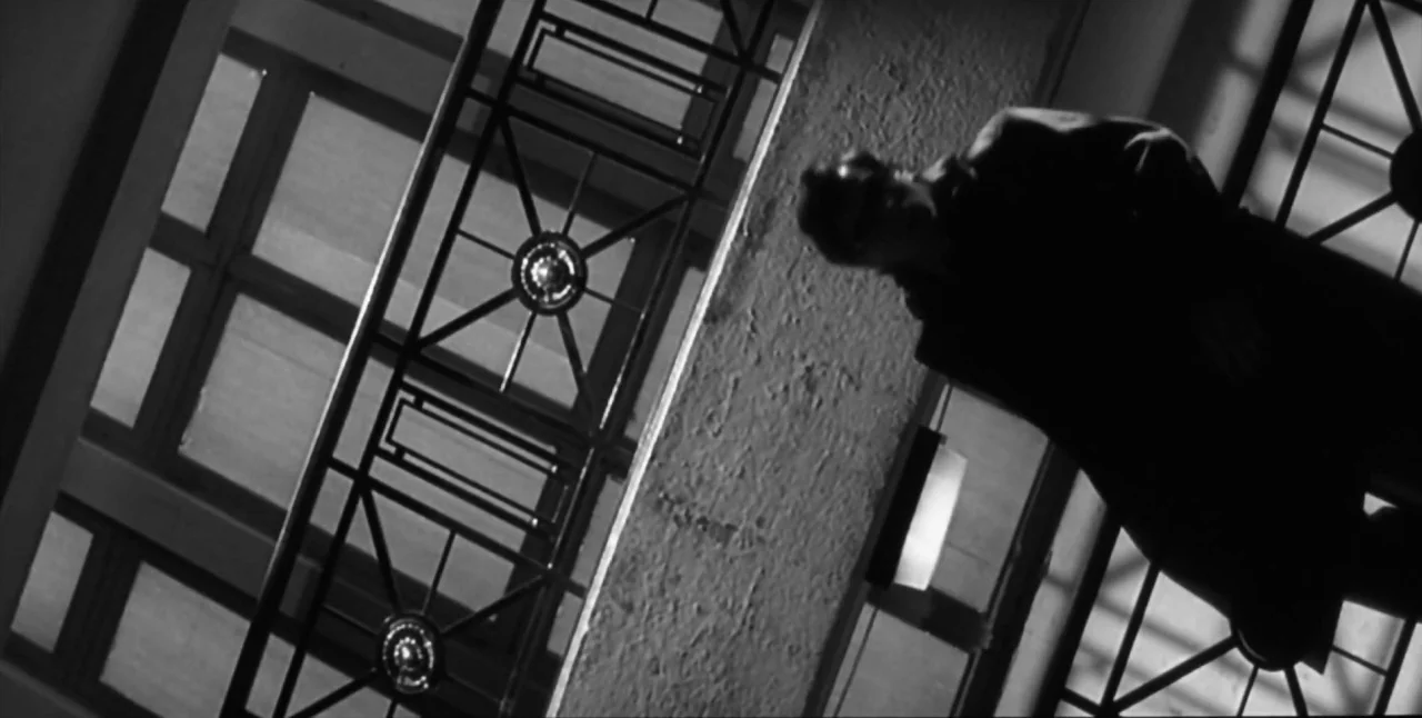 The Aerial (2007) movie still: medium — A dark staircase interior features a patterned metal railing with geometric Art Deco details. A man in a…