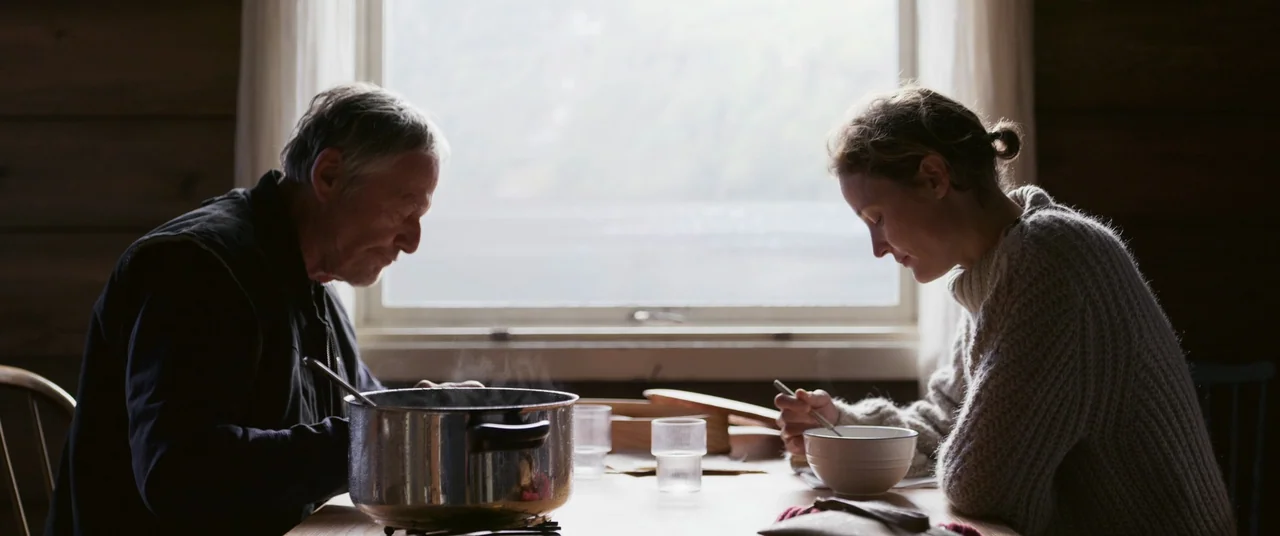 More Than Ever (2022) movie still: medium — A wooden dining table sits in front of a window overlooking a landscape. An older man and a woman sit…