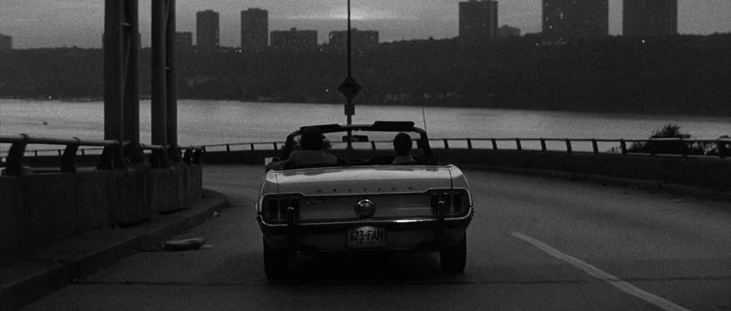 Manhattan (1979) movie still: wide — A wide shot shows a convertible driving on a bridge during dusk. The city skyline is visible in the background…