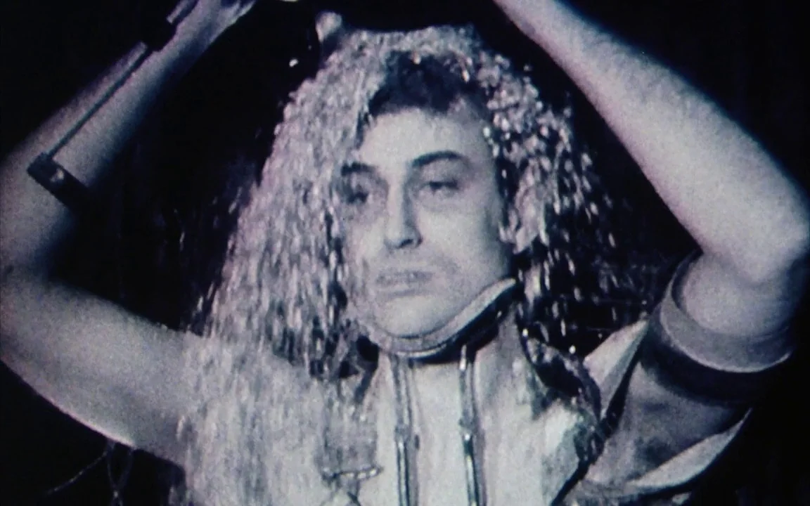 The Last of England (1987) movie still: closeup — A man stands in a dark environment with water streaming down over his head. He wears a metal neck…