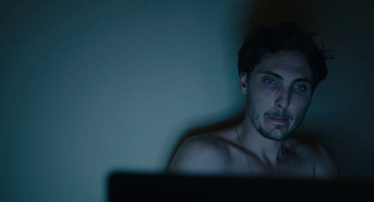 Lingua Franca (2019) movie still: closeup — A dark, minimally furnished space illuminated by an off-screen light source. A shirtless man sits facing…