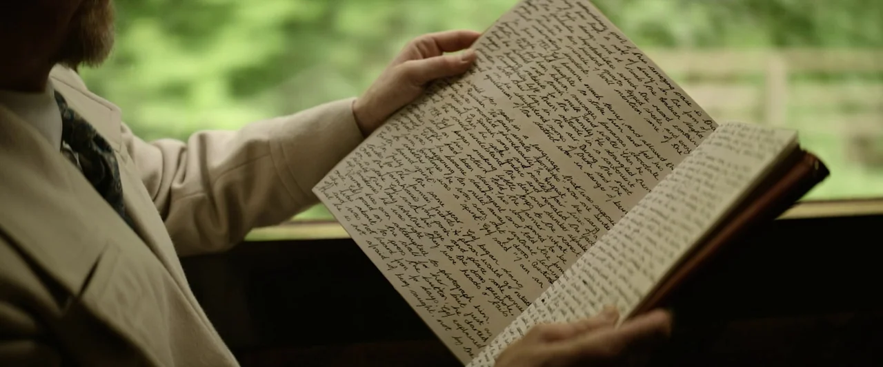 Nandor Fodor and the Talking Mongoose (2023) movie still: closeup — A man holds a notebook open, revealing dense handwritten text on aged paper. The…
