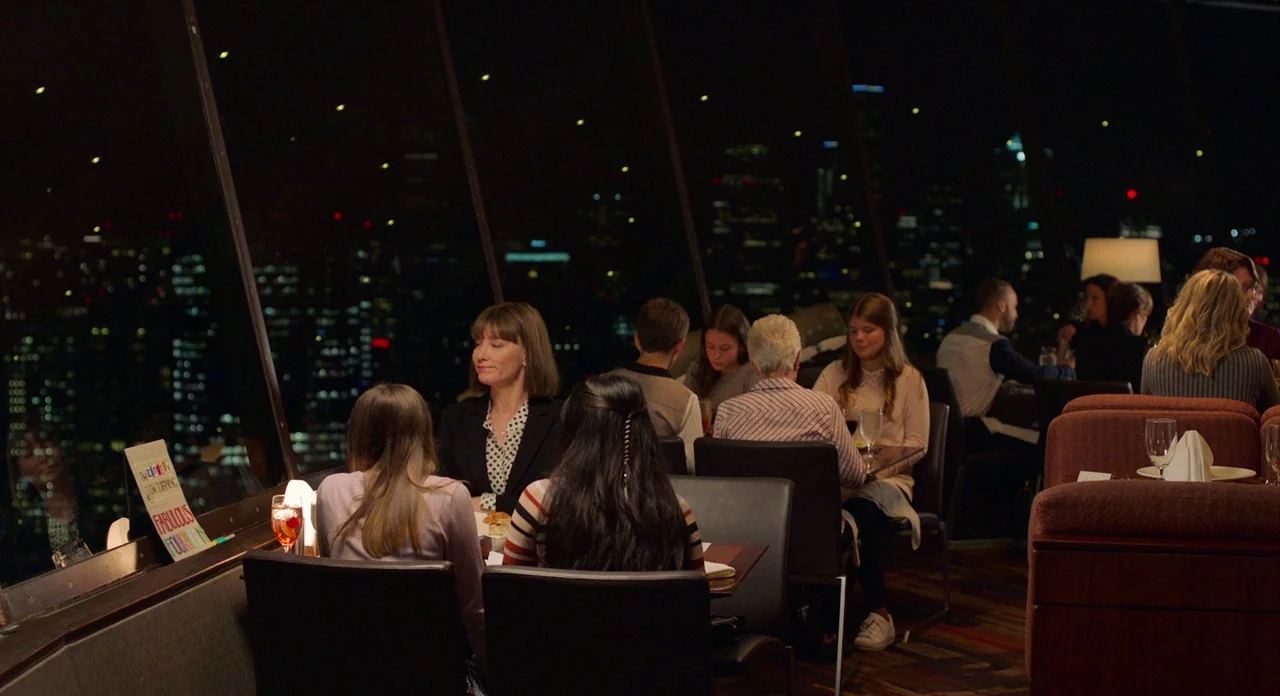 Where’d You Go Bernadette (2019) movie still: wide — A high-floor dining space features large slanted windows overlooking a sprawling nighttime city…