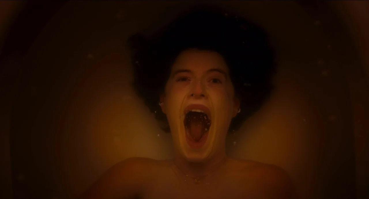 Men (2022) movie still: closeup — A dark, amber-lit bathtub contains water with suspended air bubbles. A woman is submerged with her face pointed…