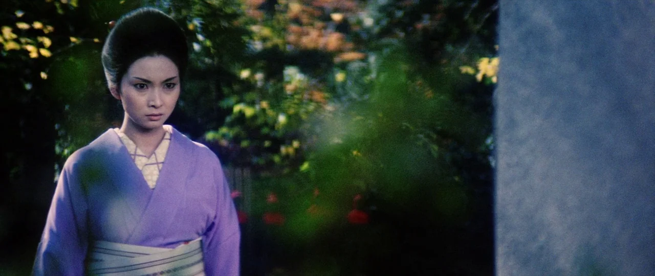 Lady Snowblood (1973) movie still: medium — A Japanese woman in a traditional kimono stands outdoors among dense, green foliage. She gazes…