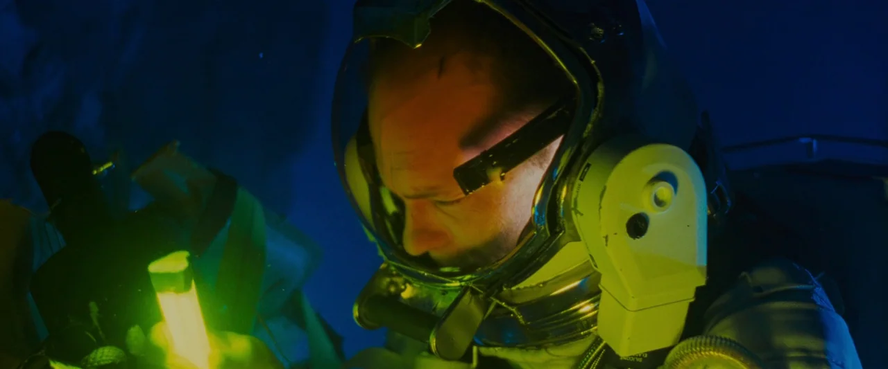 The Abyss (1989) movie still: closeup — A white pressurized space suit helmet is illuminated from below in the dark. An astronaut peers down at a…