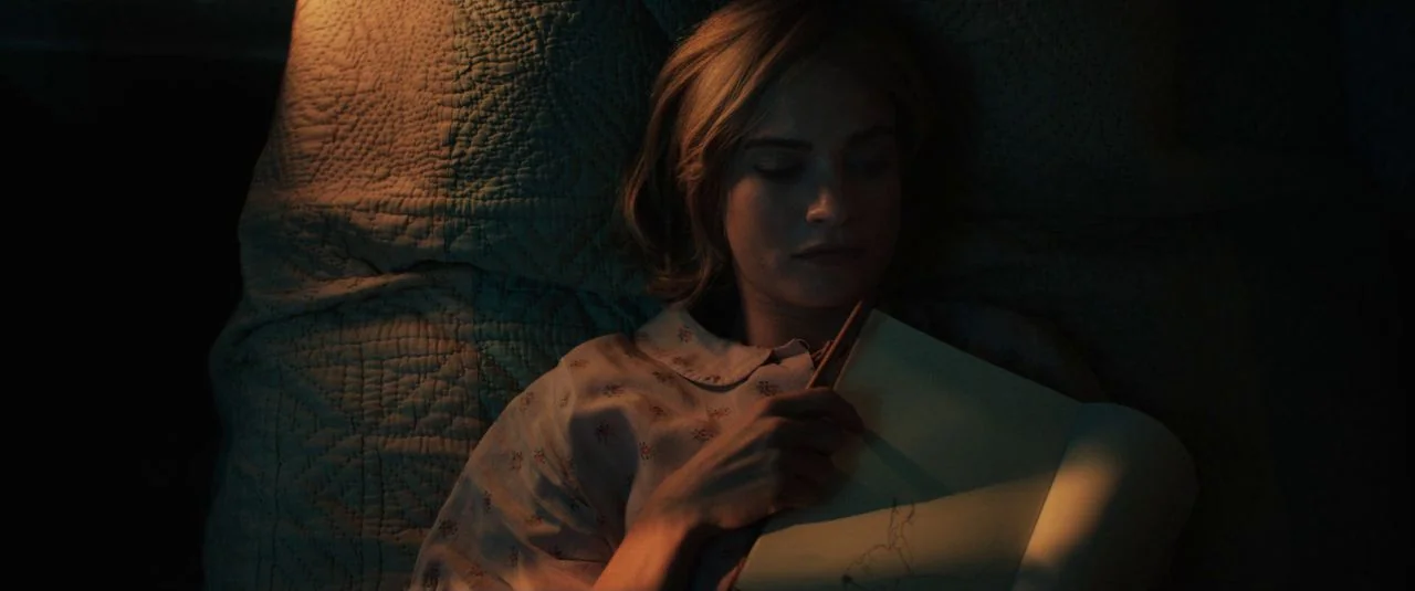 Rebecca movie still: closeup — A woman lies in bed at night with a patterned quilt in the background. She holds a pencil and sketchbook while…