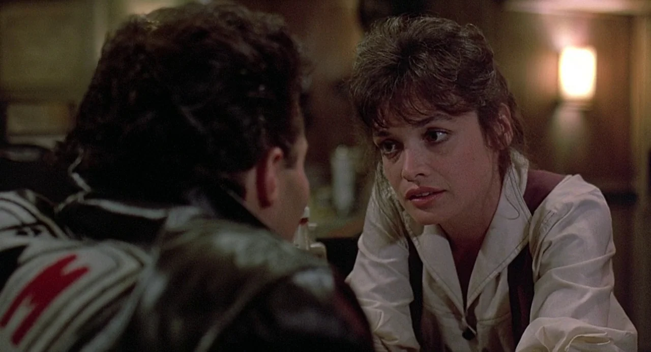 Streets of Fire (1984) movie still: medium — A dark corner of a diner with paneled walls and a dim wall lamp. A man in a leather jacket sits across…