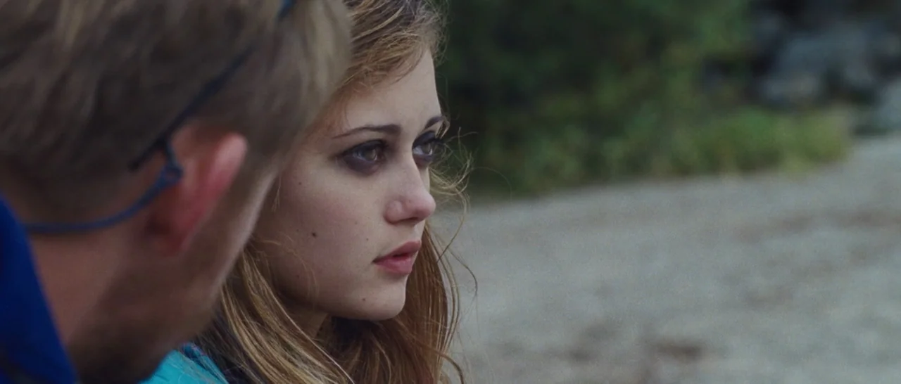 Wildlike (2014) movie still: medium — A young woman sits on a beach with a blurred man in the foreground. They appear to be engaged in a quiet…