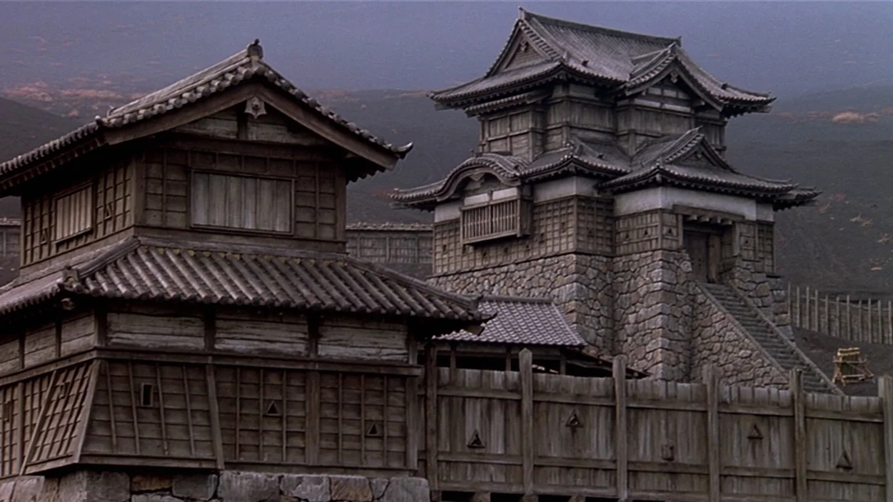 Ran (1985) movie still: establishing — Traditional Japanese wooden castle buildings sit behind a large wooden palisade wall. A stone-walled…