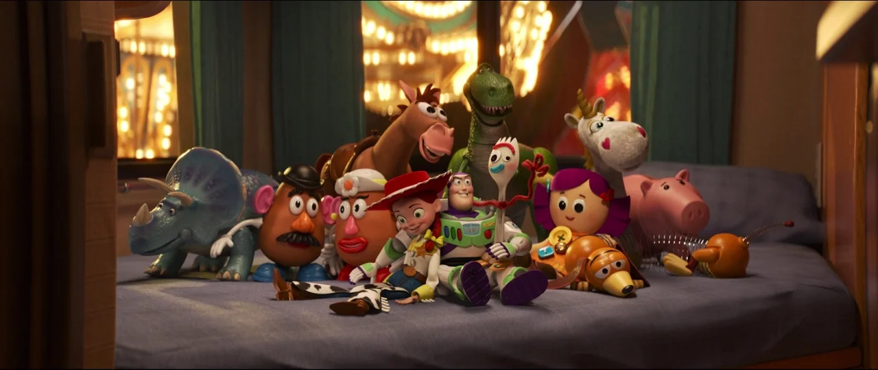 Toy Story 4 (2019) movie still: wide — A collection of colorful plastic and plush toys sits on a blue bedspread. The background shows blurred warm…