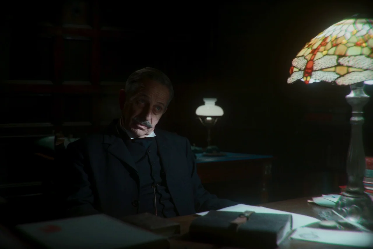 The Settlers (2023) movie still: medium — A dimly lit study contains a wooden desk with books and a glass lamp. An older man sits at the desk while…