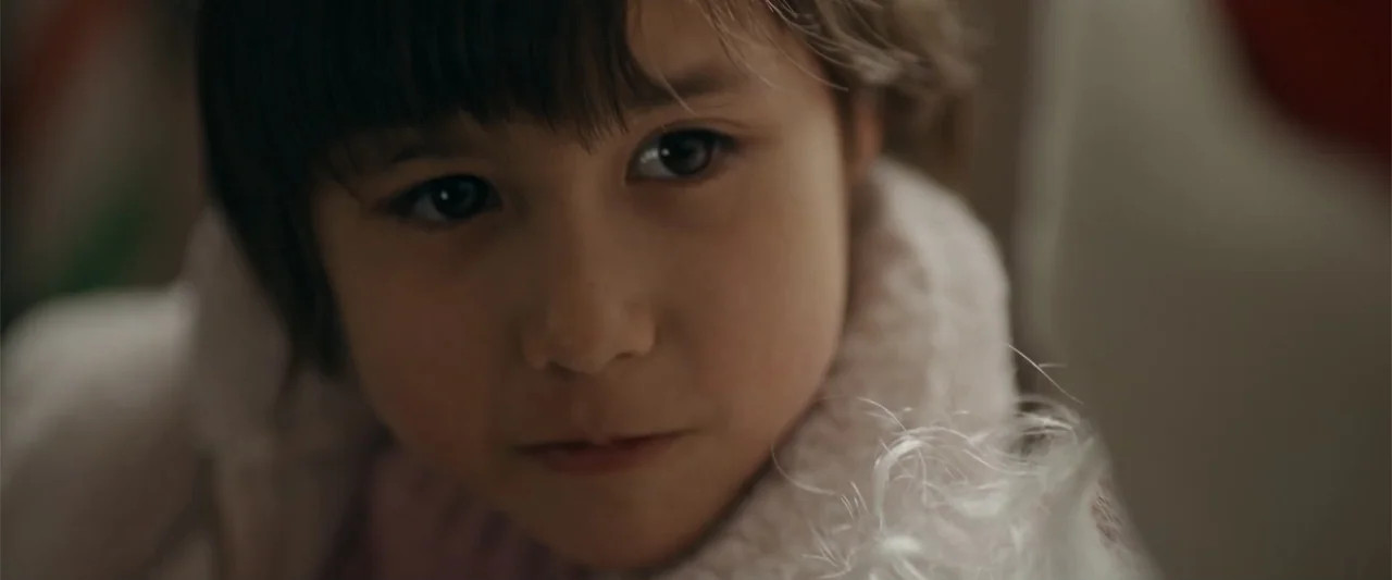 Silent Night, Deadly Night (2025) movie still: closeup — A close-up view of a child in a dimly lit indoor space. A young girl with dark hair looks…