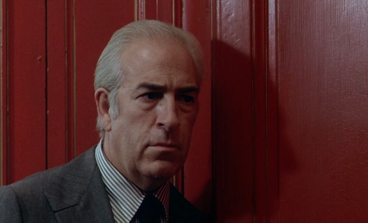 The Beast (1975) movie still: closeup — An older man stands against a painted wooden wall with vertical paneling. The bright red wall serves as the…