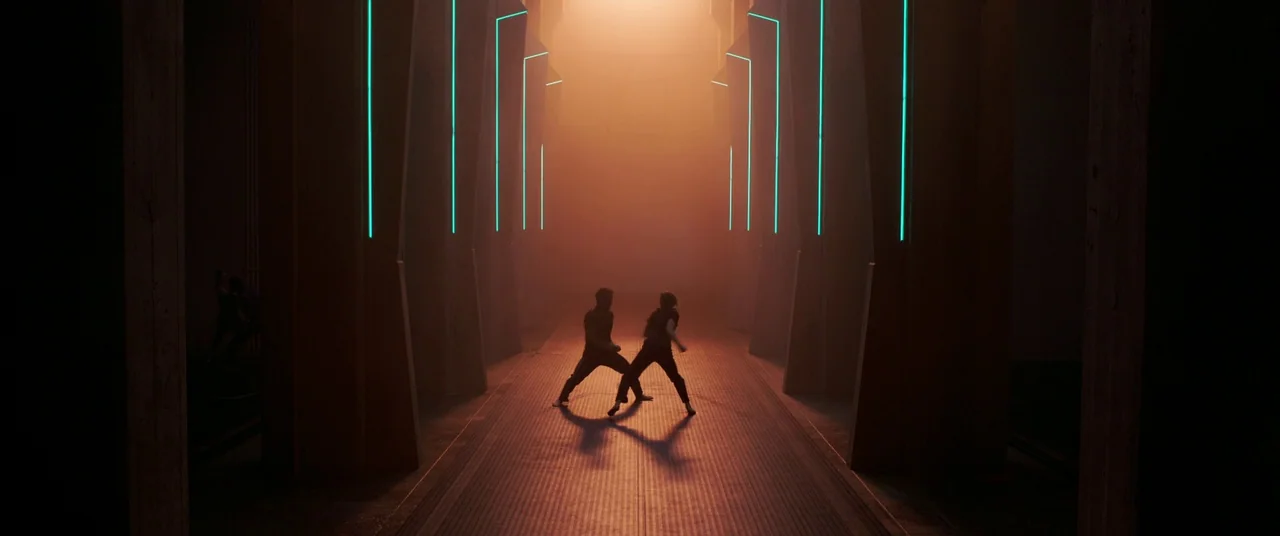 Captain Marvel (2019) movie still: wide — A long symmetrical hallway features tall concrete pillars accented with vertical glowing neon cyan strips.…