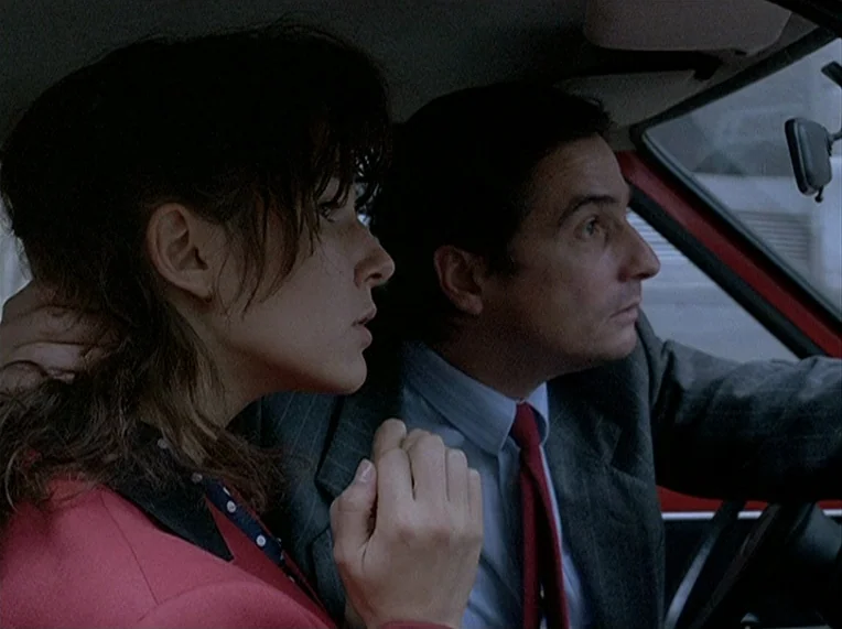 Detective (1985) movie still: medium — A view inside a vehicle cabin with two individuals positioned in the front seats. The man operates the…