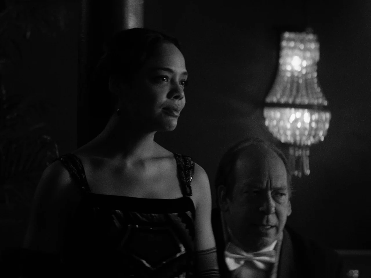 Passing (2021) movie still: medium — A dark, elegant interior space features a glowing crystal wall sconce in the background. A woman and an older…