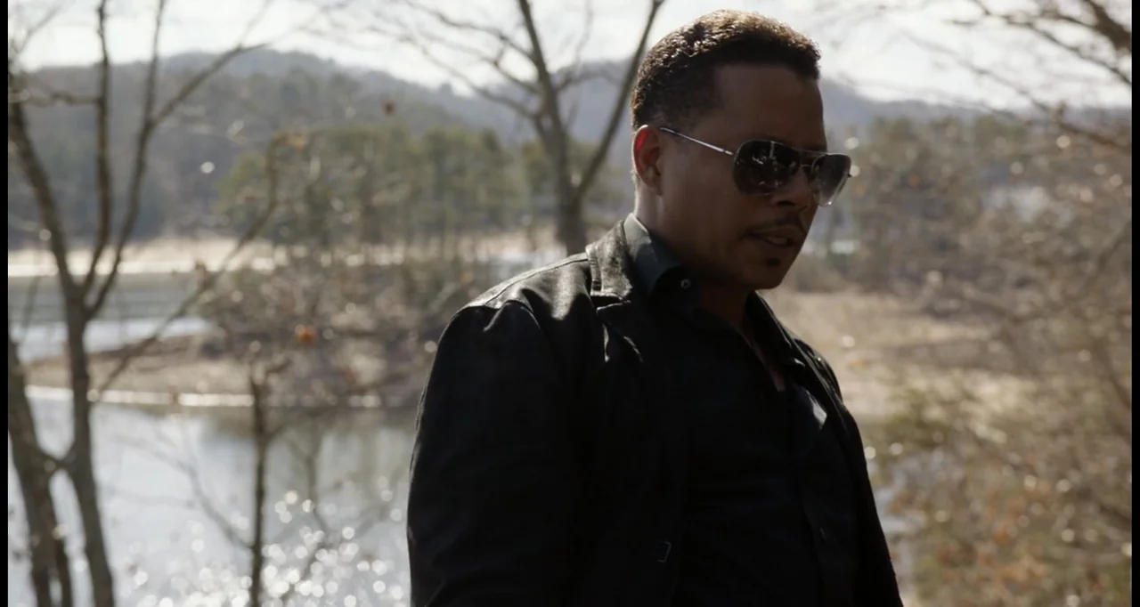 Sabotage (2014) movie still: medium — A man stands outdoors near a calm lake with leafless trees in the background. The man wears a black leather…